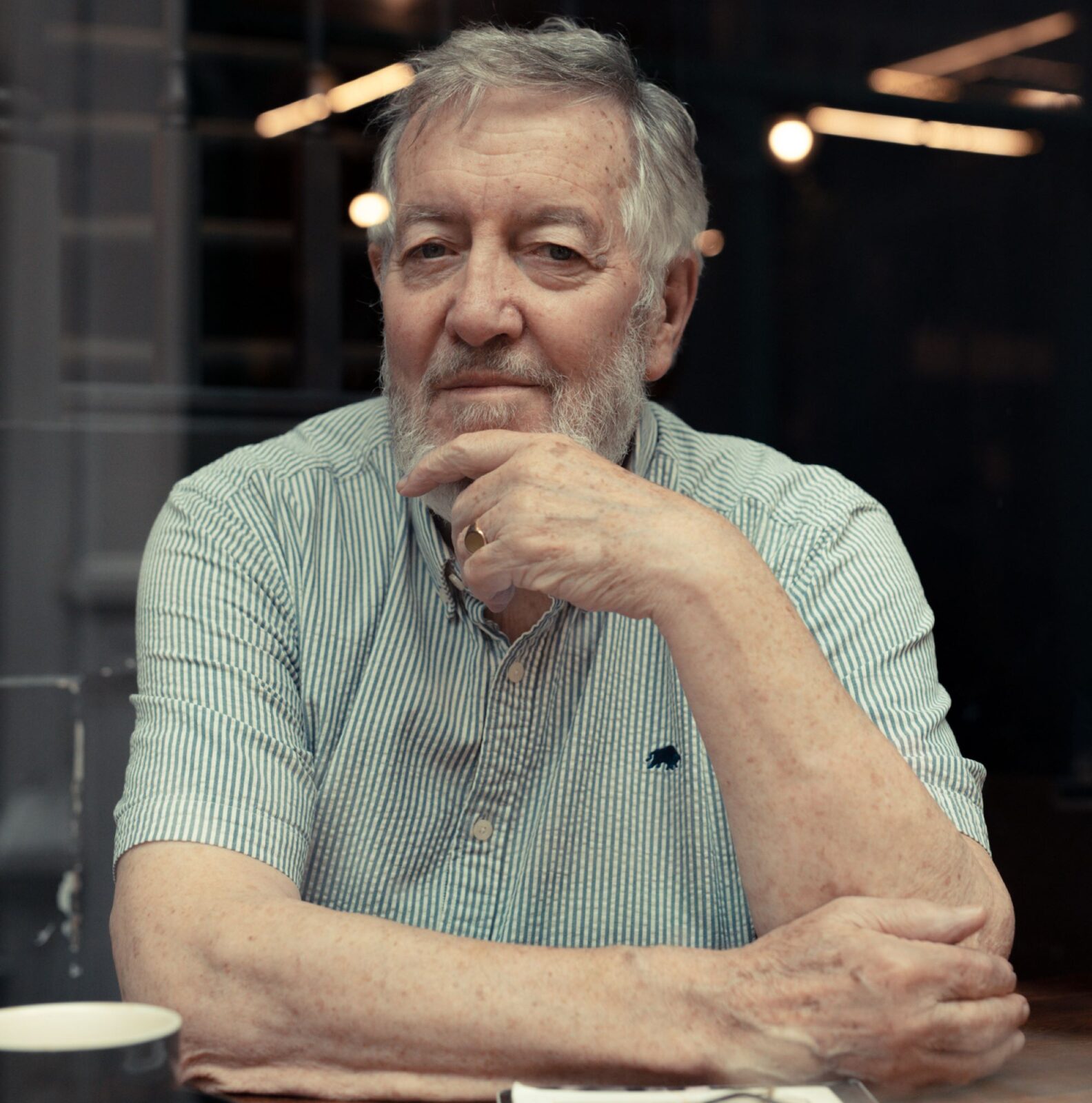 Hywel Gwynfryn. An older white man with thick grey hair and beard wearing a green and white short sleeved, pin-striped shirt.